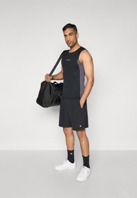 Schwarzes Sporttanktop mit grauem seitlichem Einsatz; schwarze Shorts; weiße Sneakers; trägt eine schwarze Sporttasche; Logo auf Tanktop und Shorts.