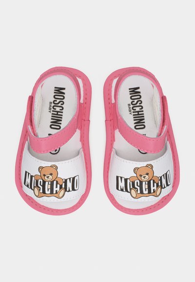 Παιδικά σανδάλια με ροζ λουράκια, λευκό μπροστινό μέρος και λογότυπο αρκουδάκι με τη σήμανση "Moschino" σε κάθε παπούτσι.