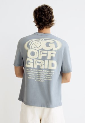 Persoană cu păr scurt și creț, purtând un tricou gri cu text mare "OFF GRID" și un paragraf imprimat pe spate, stând într-un fundal simplu.