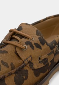 Mocassins de camurça com estampa de leopardo castanha e preta, com atacadores de pele em tom areia e ilhós de metal, sobre um fundo claro.
