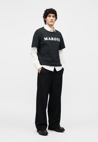 Νέος άνδρας με σγουρά μαλλιά φοράει μαύρο μπλουζάκι "MARGIE" πάνω από λευκό πουκάμισο με κουμπιά, μαύρο παντελόνι με φαρδιά μπατζάκια και σκούρα παπούτσια, στέκεται με τα χέρια στις τσέπες.