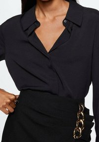 Sort langærmet skjorte med krave og to synlige knapper, parret med en højlivet nederdel med gyldne kæde detaljer.