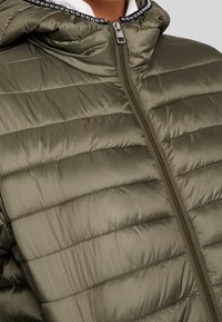 Olivově zelená péřová bunda s horizontálním prošíváním, vysokým límcem a předním zipem. Má lem s akcentem loga kolem kapuce.