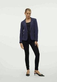 Femme portant un blazer navy, un top noir, un pantalon slim noir et des escarpins noirs à talons slingback, se tenant devant un fond clair.