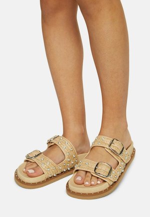 Beige slide sandals with dual silver buckle straps and decorative rhinestones. Features a textured upper and a cushioned sole with studs.
