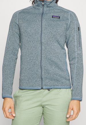 Person trägt eine hellblaue Fleecejacke mit durchgehendem Reißverschluss und einem Patagonia-Logo auf der Brust sowie hellgrüne Hose.