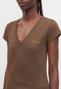 Femme portant un T-shirt marron à col en V avec manches courtes et petit texte jaune brodé sur la poitrine.