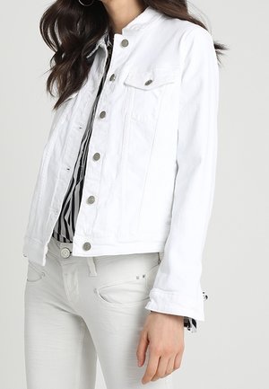 Vrouw die een witte button-down spijkerjas draagt over een zwart-wit gestreept shirt en witte broek, staande tegen een effen achtergrond.