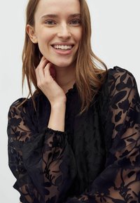 Bluză neagră din material transparent, cu un model floral, mâneci texturate și manșete gatherite. Modelul prezintă un prim-plan, evidențiind detaliile elaborate ale designului.