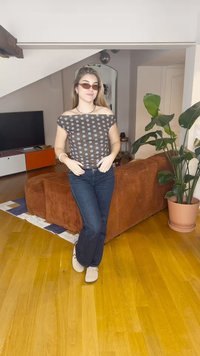 Brūna off-the-shoulder stila blūze ar zilām polka punktu rakstu, kombinēta ar tumšām džinsa biksēm un gaišām kurpēm, atrodoties modernā viesistabā ar augiem.