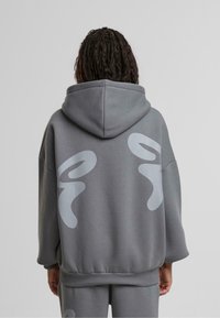 Sudadera gris de gran tamaño con un gran diseño abstracto en gris claro en la parte posterior, que cuenta con capucha ajustable y puños acanalados. Textura suave.