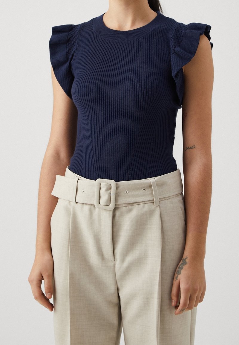 Kvinna som bär en marinblå ribbad ärmlös topp med volangärmar och högmidjade beige byxor med en bred spänne.