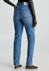 Jasnoniebieskie dżinsy z denimu o prostych nogawkach, wysokim stanie i widocznym przeszyciu. Posiadają tylne kieszenie oraz metkę "Calvin Klein" na pasie.