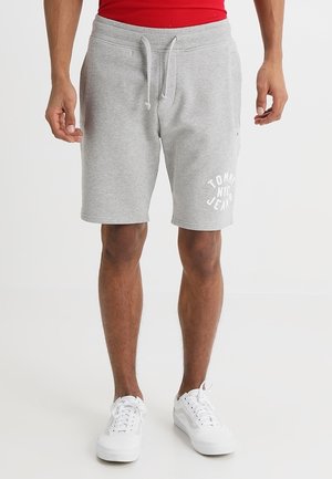 Hombre con pantalones cortos grises claros con cordón y texto "Tommy NYC Jeans" en la pierna izquierda, y zapatillas blancas, de pie sobre fondo blanco.