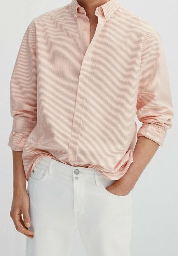 Homme portant une chemise rose clair à manches longues, boutonnée avec des fines rayures verticales, manches retroussées, et un pantalon blanc, main dans la poche.