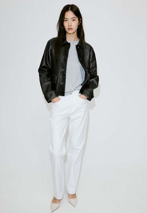 Mujer con cabello largo y oscuro que lleva chaqueta de cuero negra, camisa gris, pantalones blancos y zapatos puntiagudos beige, de pie frente a un fondo blanco.