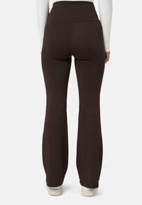 Osoba zobrazená zezadu, která má na sobě tmavě hnědé kalhoty s vysokým pasem a rozšířenými nohavicemi a bílé tenisky, na jednobarevném pozadí.