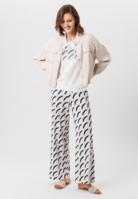 Lichtbeige jack met zilveren knopen, witte grafische t-shirt en brede broek met een zwart golvend patroon. Beige instappers.
