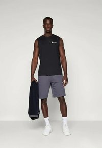 Camisa sin mangas negra con logo en el pecho, pantalones cortos grises, sosteniendo una chaqueta oscura, usando zapatillas blancas, de pie contra un fondo neutro.