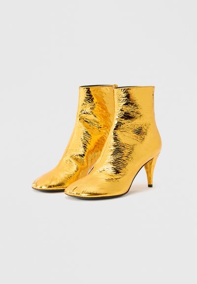 Κίτρινα μεταλλικά μποτάκια αστραγάλου με γυαλιστερή, υφή επιφάνεια, μυτερές μύτες και λεπτό, ψηλό τακούνι. Κομψός και εφαρμοστός σχεδιασμός.