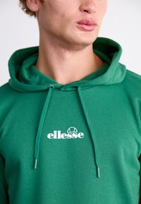 Sudadera verde con tejido texturizado, cuenta con capucha ajustable y un logo blanco de "ellesse" en el pecho. Diseño sencillo, ajuste casual.