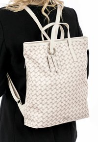Sac à dos fabriqué en tissu texturé beige avec un motif géométrique, doté de bretelles crème et d'une étiquette décorative attachée par un anneau en argent.