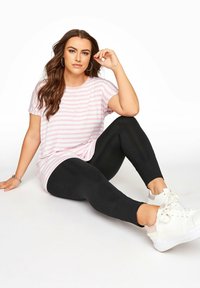 Ett ljusrosa och vitt randigt t-shirt med korta ärmar, parat med svarta leggings och vita sneakers, i sittande position mot en vit bakgrund.