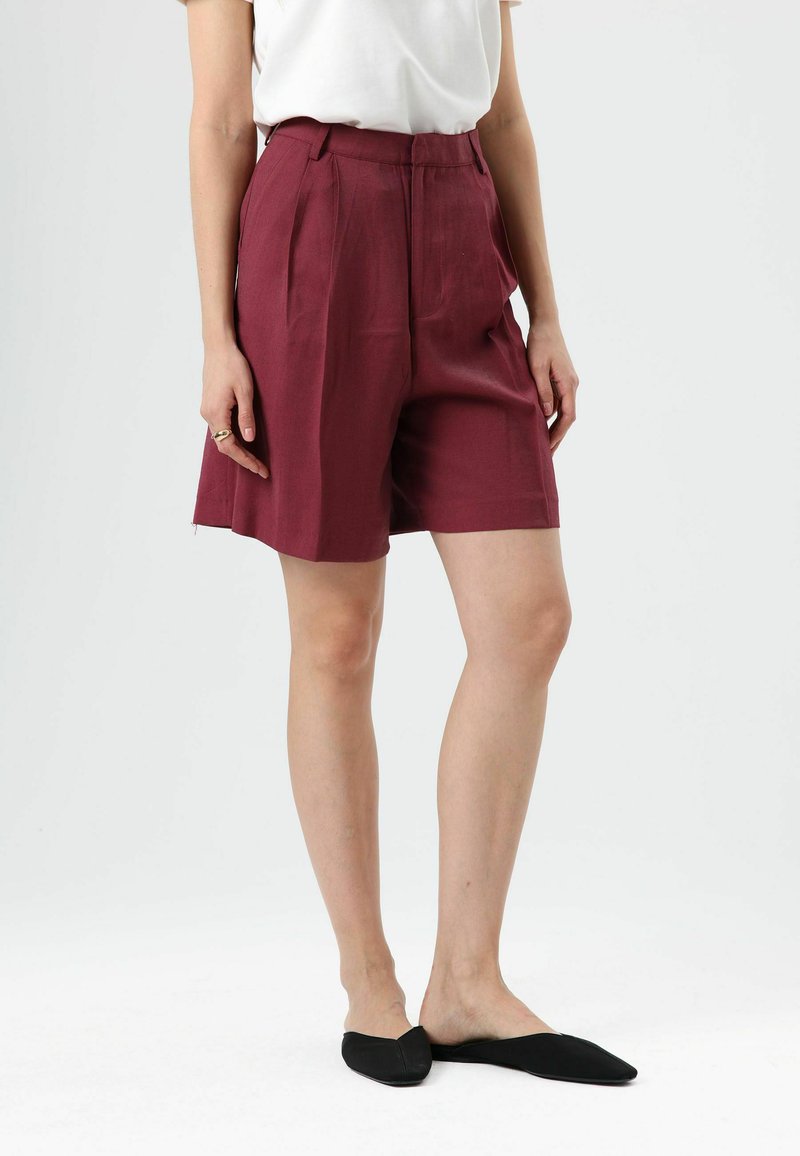 Bordeauxrode, op maat gemaakte korte broek met plooien, hoge taille en een gladde textuur. Gecombineerd met zwarte instapschoenen. Effen witte top.