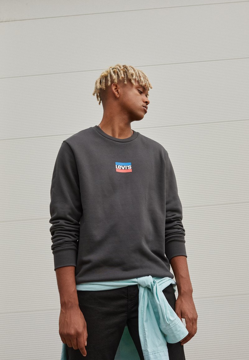 Sudadera gris con un pequeño logo de Levi's en rojo y azul, combinada con jeans negros y una camisa azul clara atada a la cintura.