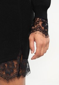 Svart stickad plagg med genomskinliga svarta spetsärmar med blommönster och fransdetaljer. Mjuk textur med en avslappnad passform.