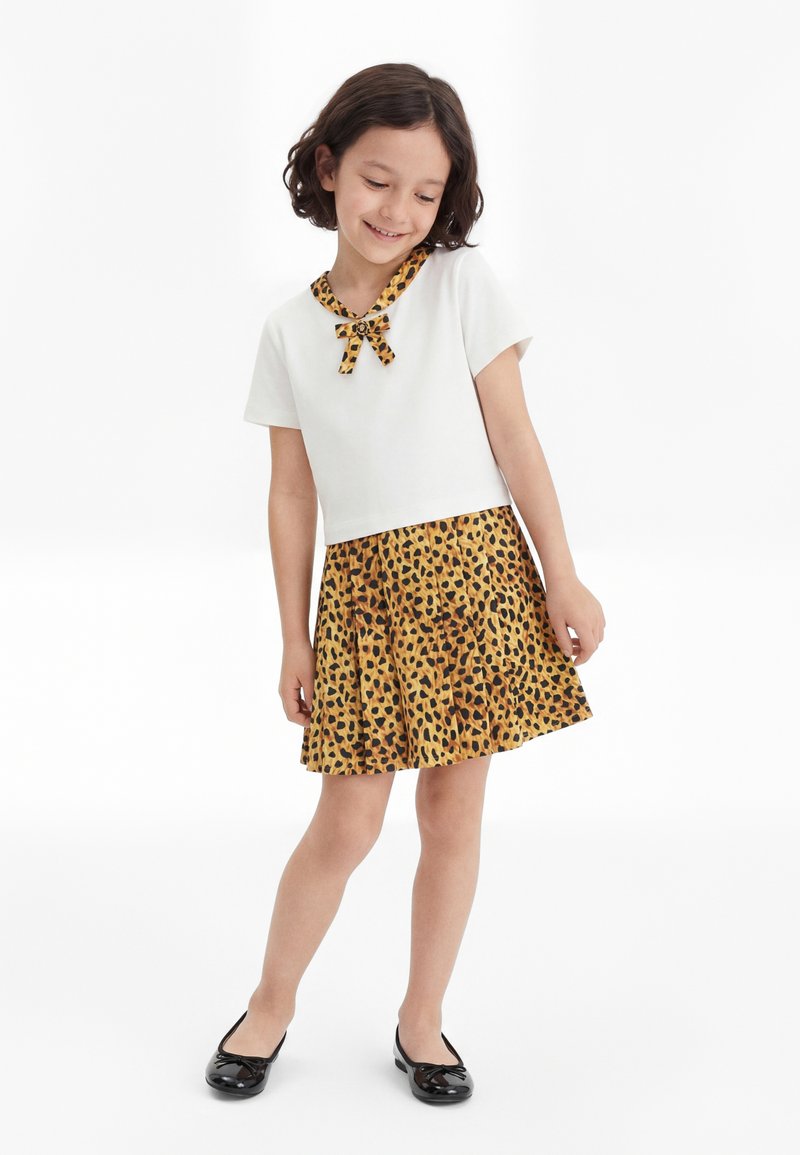 Fille portant un haut blanc à manches courtes avec un col et un nœud imprimés léopard, une jupe assortie imprimée léopard et des ballerines noires, debout et souriante.