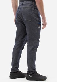 Jeans w ciemnoniebieskim odcieniu, z zwężającymi się nogawkami, wzmocnionymi szwami i bocznymi kieszeniami. W połączeniu z czarnymi butami trekkingowymi.