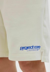 Béžové bavlnené šortky s modrým logom "project core" pri leme. Majú voľný strih a bočné rozparky na nohách pre väčšiu pohyblivosť.