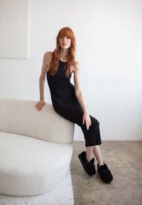 Čierne bezrukávové šaty, jemná textura, s priliehavým strihom. Modelka sedí na zaoblenom bielom syntetickom kožuchu proti svetlému pozadiu.