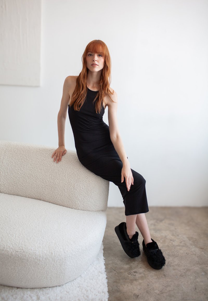 Čierne bezrukávové šaty, jemná textura, s priliehavým strihom. Modelka sedí na zaoblenom bielom syntetickom kožuchu proti svetlému pozadiu.