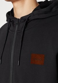 Felpa nera con zip e cordini, caratterizzata da un'etichetta marrone testurizzata con la scritta “HUGO” ricamata sul petto sinistro. Tessuto morbido.