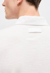 Homme portant une chemise blanche à col, vu de dos, mettant l'accent sur la zone du col de la chemise et une petite boucle en tissu sur le col.