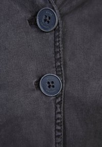 Ciemnoszary materiał o gładkiej fakturze ma dwa granatowe guziki, każdy z czterema otworami, oraz subtelne detale przyszycia wzdłuż brzegów.