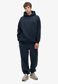 Sudadera de algodón azul marino con bolsillo canguro, combinada con joggers a juego. Cuenta con un ajuste relajado y puños de canalé. Llevada con zapatillas grises.