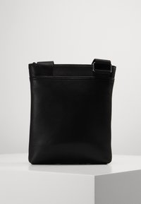 Černá kožená křížová taška s nastavitelným popruhem vystavená na bílém podstavci na jednoduchém světle šedém pozadí.