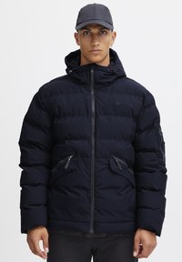 Pufjakke i marineblå med lynlås, justerbar hætte, to forreste lynlåslommer og struktureret stof. Letvægts- og isoleret design.