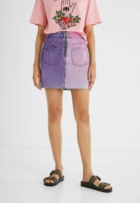 Minifalda de mezclilla con un degradado de púrpura a rosa, que cuenta con dos bolsillos traseros y un cierre de cremallera en la parte delantera. Combinada con una camiseta gráfica rosa.