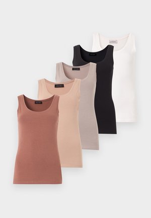Kvinde står mod hvid baggrund iført hvid ærmeløs top, røde bukser med vide ben, brune sandaler og gyldne creoløreringe.