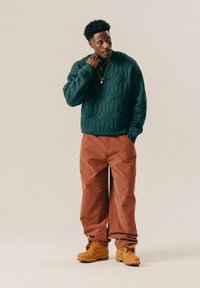 Grüner gesteppter Sweatshirt mit erhabenem Muster, rote karierten weite Hosen und braune Stiefel mit Schnürsenkeln und robusten Sohlen. Goldene Halskette als Akzent.