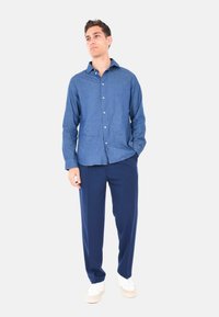 Camicia in chambray azzurro chiaro con design a bottoni e maniche lunghe, abbinata a pantaloni navy e sneakers bianche su uno sfondo neutro.