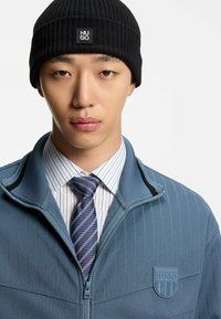 Black ribbed knit beanie with a logo, paired with a light blue zip-up jacket featuring vertical lines and an embossed logo, layered over a white striped shirt and blue-striped tie.