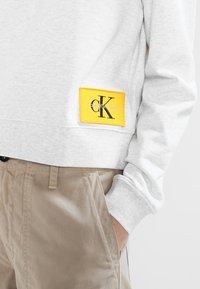 Biała bawełniana bluza z ribbowanym dołem i żółtą metką z czarnym napisem. Zestawiona z jasnobeżowymi spodniami.