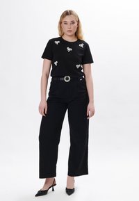 Schwarzes T-Shirt mit fünf dekorativen weißen Schleifen, kombiniert mit hoch geschnittenen schwarzen Hosen und einem schwarzen Gürtel mit einer runden silbernen Schnalle.