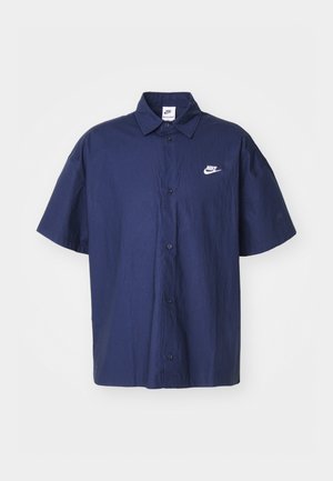 Jūras spēku īsām piedurknēm krekls ar pogām, kuram ir krakšķīga tekstūra un balts Nike logo kreisajā krūtī. Iezīmē standarta apkakli un melnas pogas.