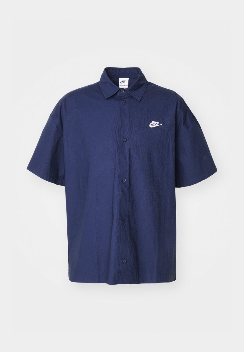 Chemise à manches courtes bleu marine à boutons avec une texture froissée et un logo Nike blanc sur la poitrine gauche. Elle présente un col classique et des boutons noirs.